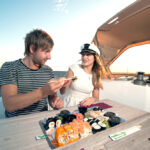 Dvojica si vychutnáva sushi na palube jachty počas plavby pri západe slnka.